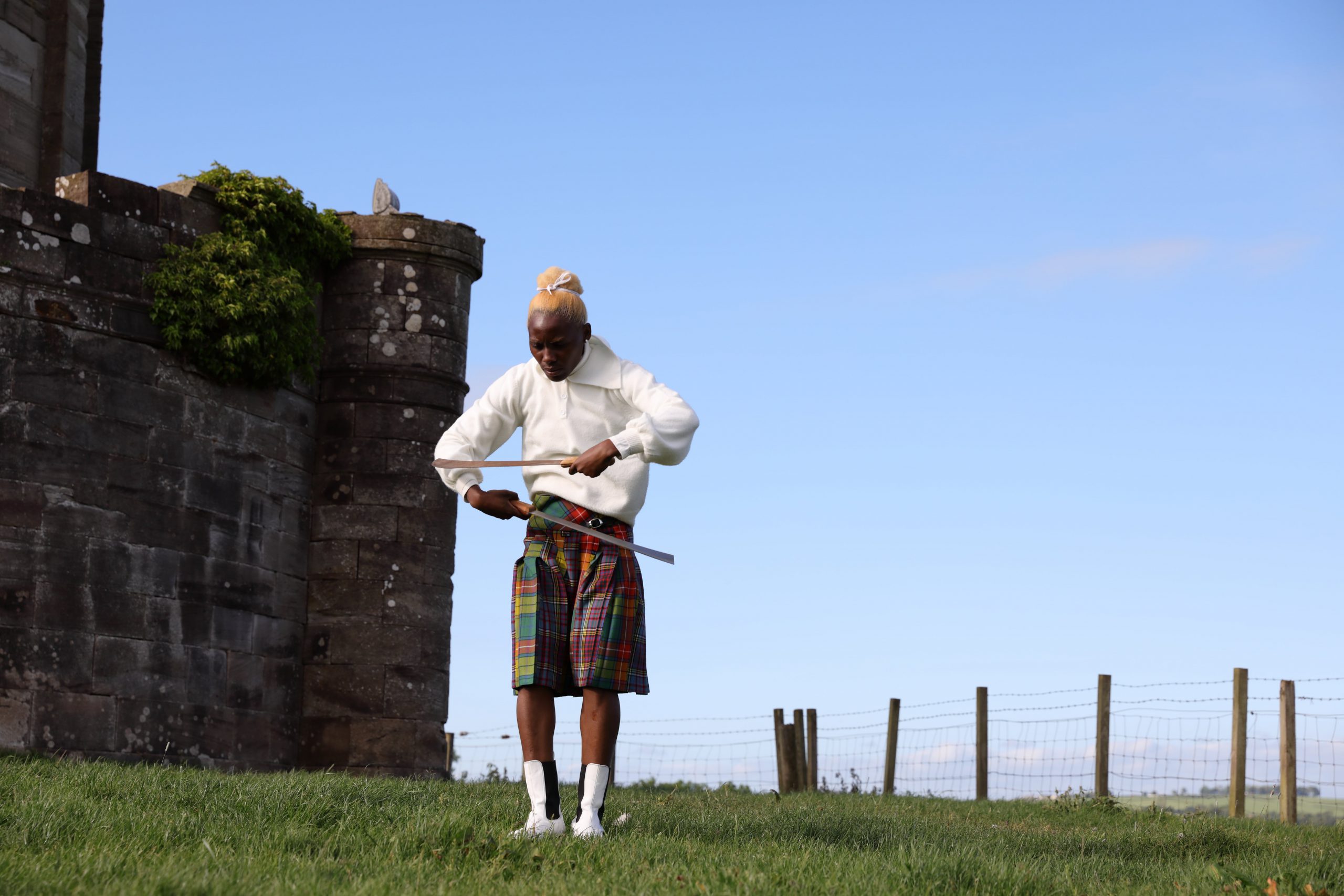 An image still from the film Lagareh of a Black dancer wearing a kilt and a white shirt. The dancer holds two long knives in her hands and stands on green grass with a blue sky behind her and a castle-like building to her left.